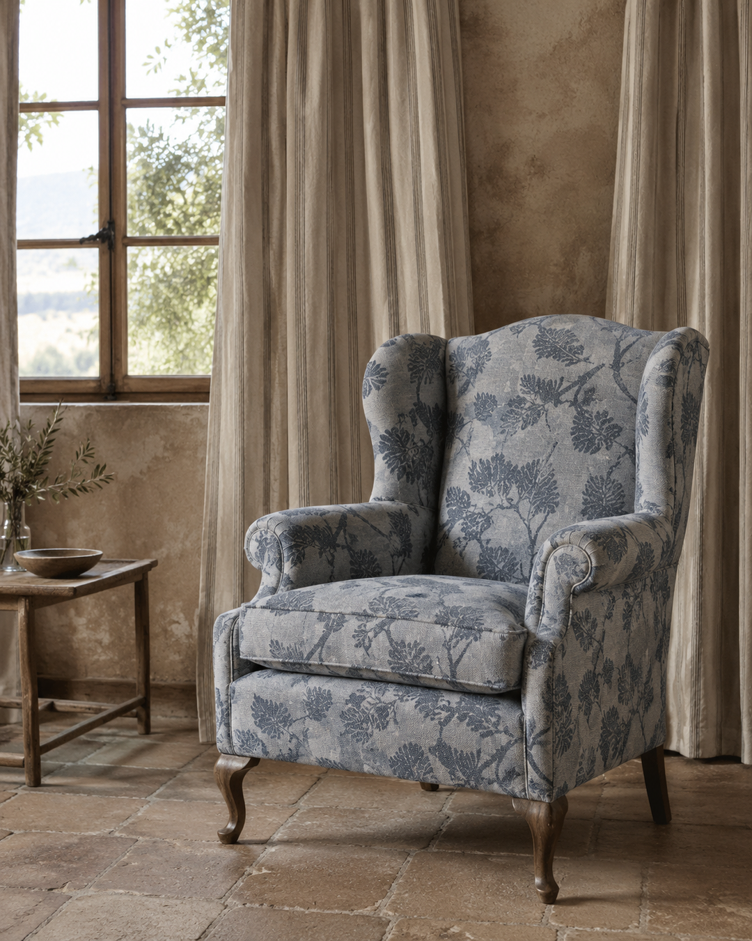 A classic wingback armchair upholstered in a blue and gray botanical floral pattern, set in a rustic room with beige curtains, a stone floor, and a wooden side table.